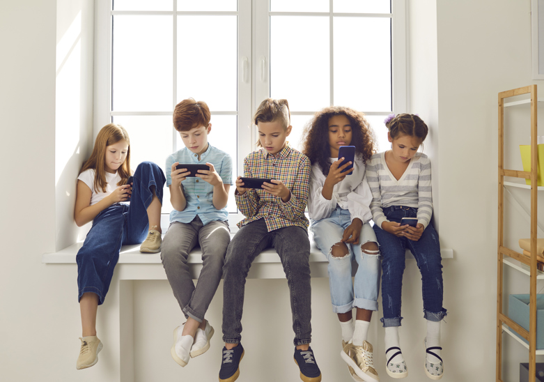 A row of children sat in front of a window, all scrolling on their mobile devices. 